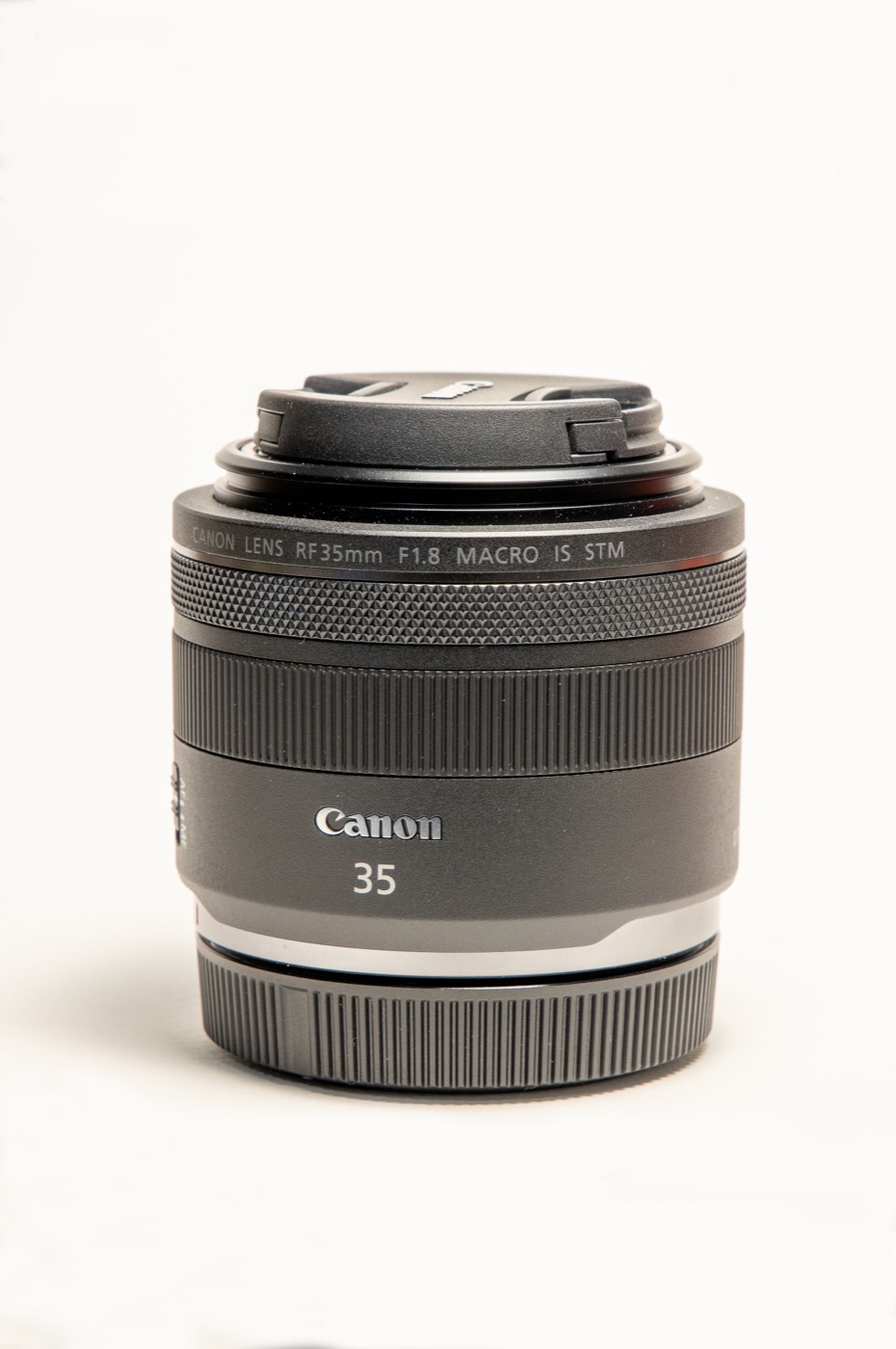 A Canon RF 35mm F1.8 Macro lens with its cap on, standing on a white background