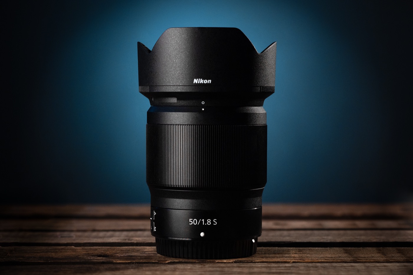 A black Nikon 50/1.8 S lens with its hood fitted, standing on dark wood against a blue background